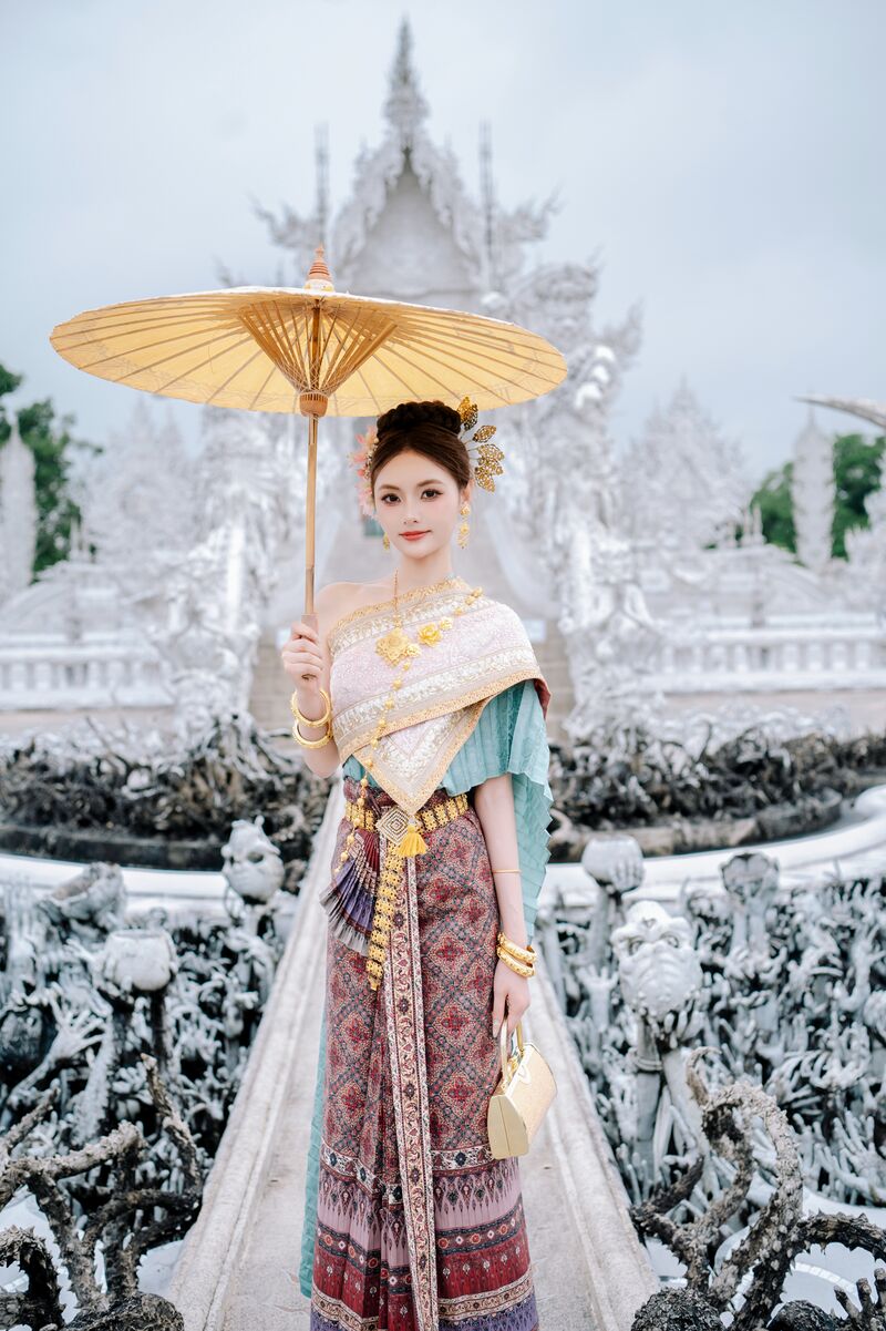 ผู้หญิงในชุดไทยถือร่มกระดาษสีทองบนสะพานไปยังวัดร่องขุ่น สองข้างเป็นประติมากรรมมือสีขาว