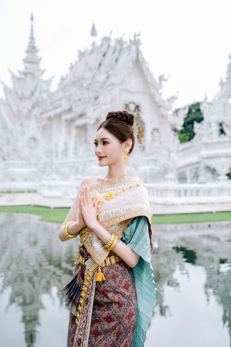 ผู้หญิงในชุดไทยพนมมือไหว้ริมสระน้ำ ด้านหลังเป็นวัดร่องขุ่นและเงาสะท้อนในน้ำ