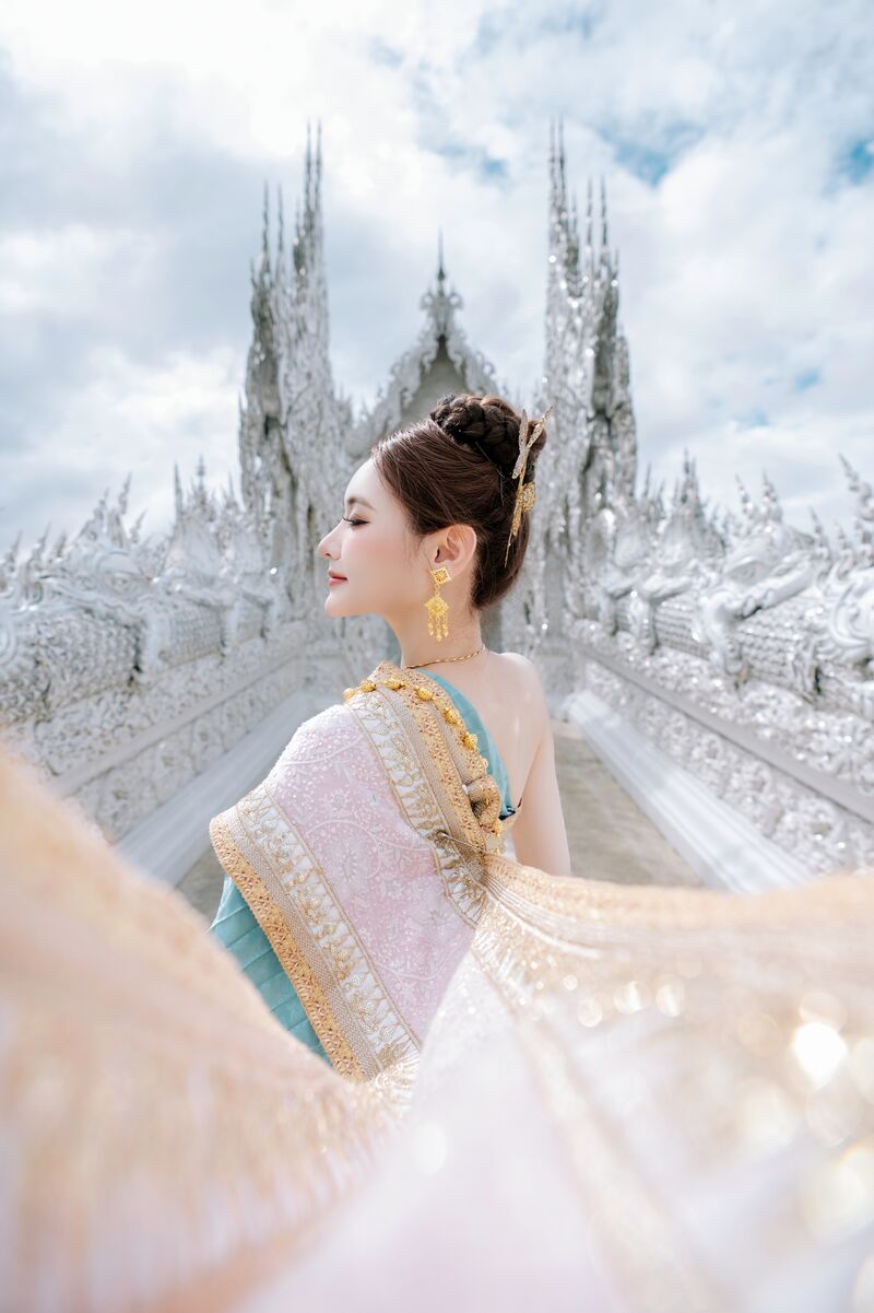 ผู้หญิงหันข้าง ผ้าสไบสีชมพูทองปลิวไสวตัดกับยอดแหลมของวัดร่องขุ่น