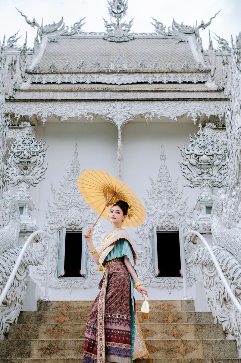 ผู้หญิงในชุดไทยถือร่มกระดาษสีทองบนบันไดหน้าอาคารสีขาวในวัด