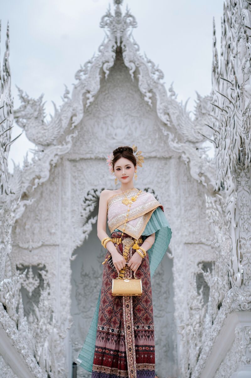 ผู้หญิงในชุดไทยถือกระเป๋าสีทอง ยืนในซุ้มประตูแกะสลักสีขาว