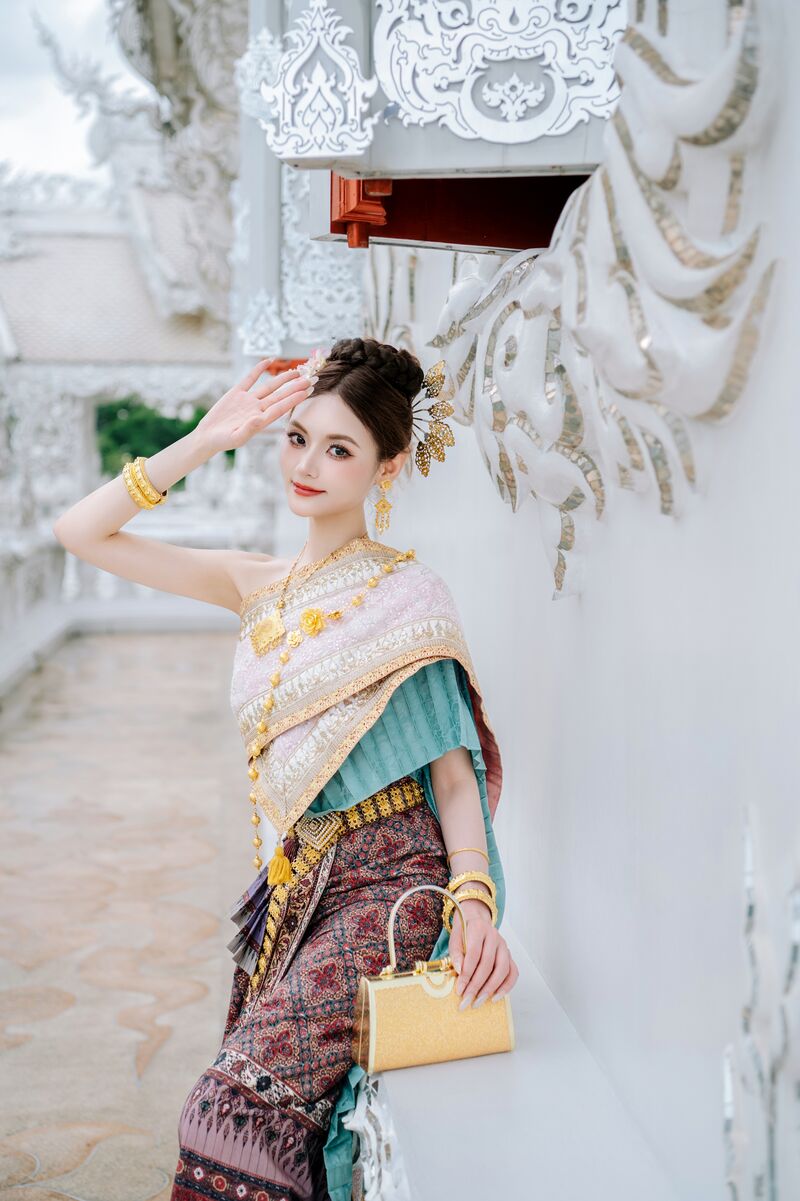 ผู้หญิงในชุดไทยยกมือขึ้นแตะหน้าผาก ข้างผนังสีขาวประดับกระจกเงิน