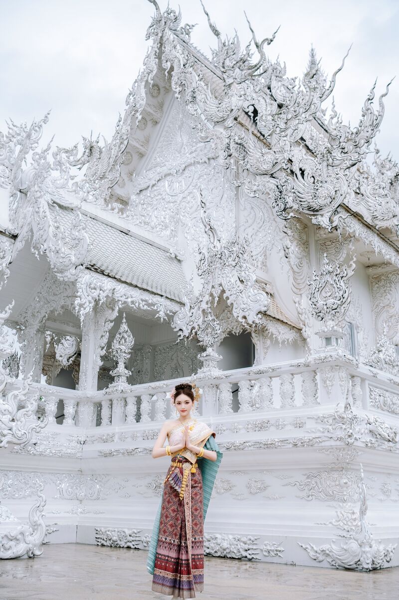 หลังคาและระเบียงแกะสลักสีขาวของวัดร่องขุ่น ด้านหน้ามีผู้หญิงในชุดไทยพนมมือไหว้