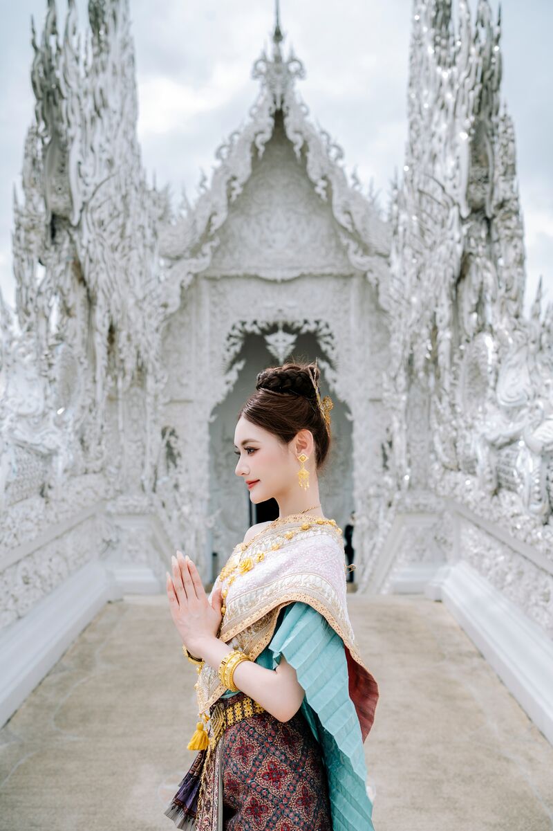 ผู้หญิงในชุดไทยพนมมือไหว้ในท่าหันข้างบนทางเดินสีขาว ด้านหลังเป็นซุ้มประตูวัด