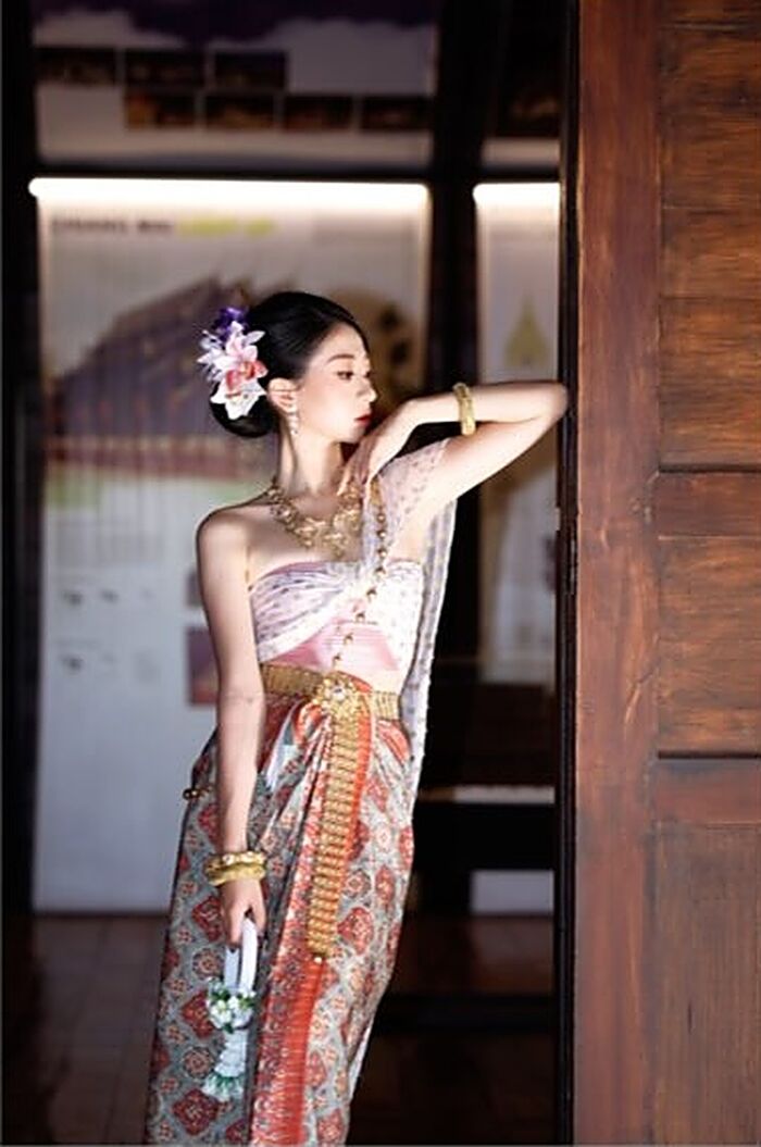 ชุดผกา: สไบสีชมพูอ่อนกับผ้านุ่งสีส้มและเขียวอมฟ้า ยืนที่ประตูไม้