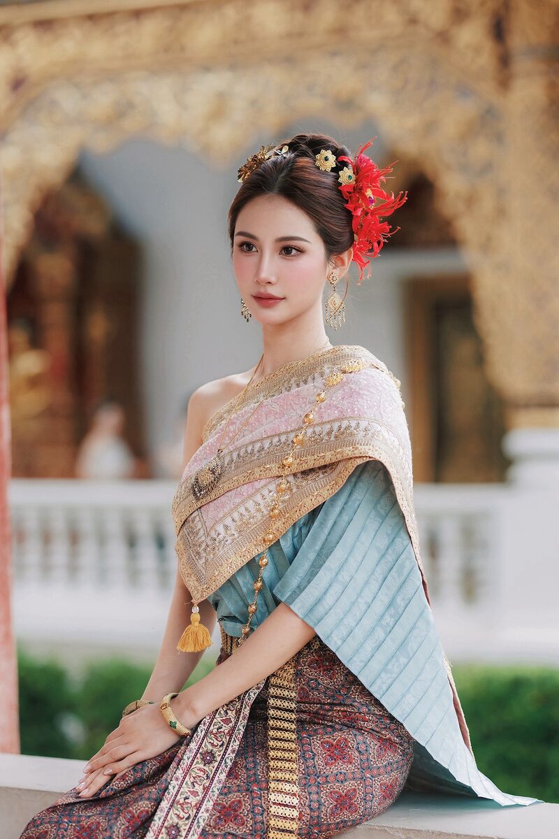 หญิงสาวห่มสไบปักทองทับผ้าจีบสีฟ้าอ่อน นั่งอยู่หน้าวิหารสีทอง