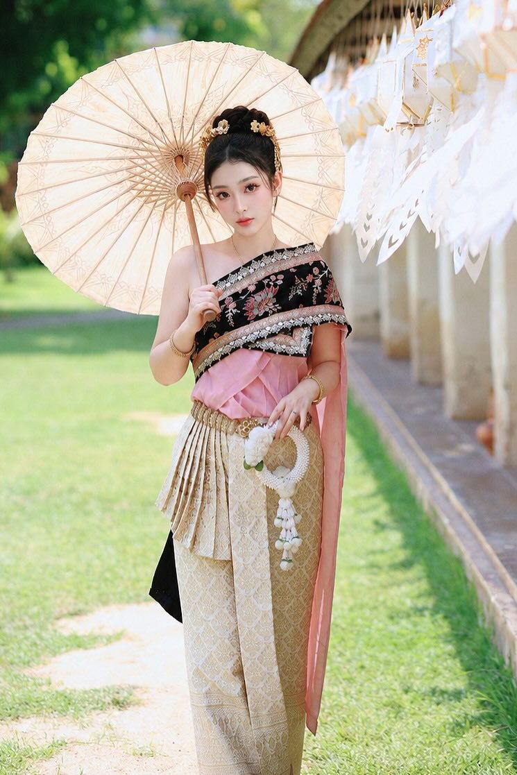 หญิงสวมชุดไทย ห่มสไบปักสีดำทับเสื้อผ้าไหมสีชมพู นุ่งผ้าจีบสีทอง ถือร่มกระดาษ ยืนบนทางเดินในสวนที่ประดับเครื่องแขวนกระดาษสีขาว