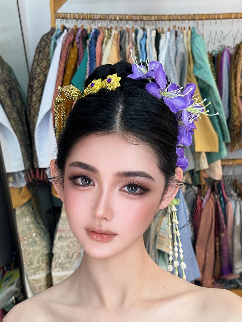 Young woman with a traditional Thai updo, purple flowers and gold hairpins, in front of a rail of costumes