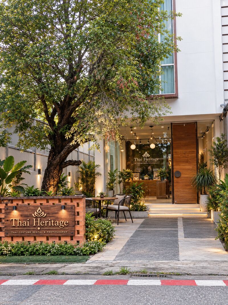หน้าร้าน Thai Heritage ยามเย็น ป้ายไม้ “Thai Heritage” บนกำแพงอิฐเตี้ย ต้นไม้ใหญ่ และหน้าร้านกระจกที่เปิดไฟสว่างกับประตูไม้บานสูง