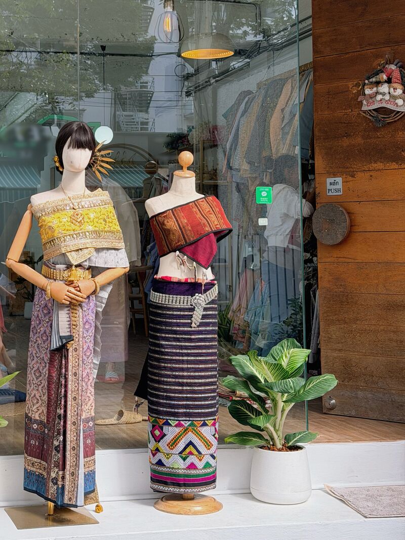 ตู้โชว์หน้าร้าน Thai Heritage หุ่นสองตัวสวมชุดไทย ตัวหนึ่งห่มสไบสีทองกับผ้านุ่งลายไหม อีกตัวสวมเสื้อลายทางกับผ้าซิ่นทอ ข้างกระถางต้นไม้และประตูไม้
