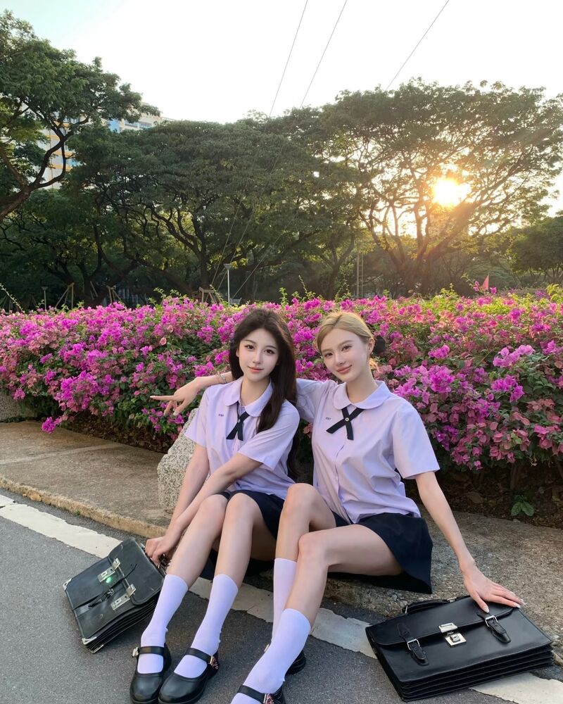 Two friends in Thai school uniforms sitting by a bougainvillea hedge at sunset