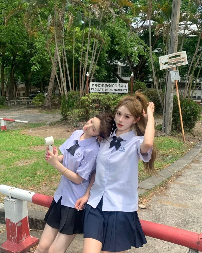 สาวสองคนในชุดนักเรียนไทยเอียงศีรษะชิดกันบนทางเดินร่มรื่นในรั้วมหาวิทยาลัย